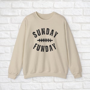 Op de afbeelding: Een beige sweatshirt met een zwarte grafische print die "SUNDAY FUN DAY" zegt met een voetbalnaadontwerp tussen de woorden.