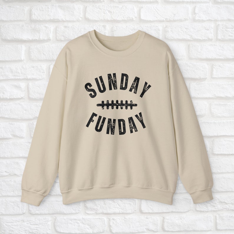 Op de afbeelding: Een beige sweatshirt met een zwarte grafische print die "SUNDAY FUN DAY" zegt met een voetbalnaadontwerp tussen de woorden.