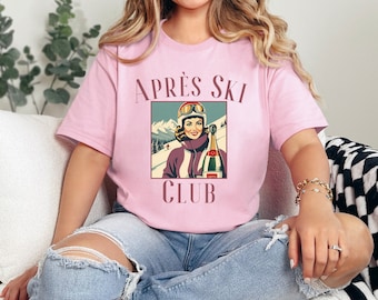 Könnte beinhalten: Ein pinkfarbenes T-Shirt mit dem Text "APRÈS SKI CLUB" und einer Vintage-Illustration einer Frau in Skiausrüstung, die eine Flasche Champagner hält.
