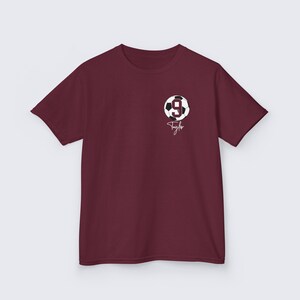 May include: Burgundy t-shirt with a white soccer ball graphic and the number 9. The name Taylor is written in white cursive below the ball.