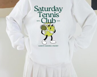 May include: White sweatshirt with a green graphic of a tennis ball with a face and the text "Saturday Tennis Club Est. 2025 Always Causing A Racket"
