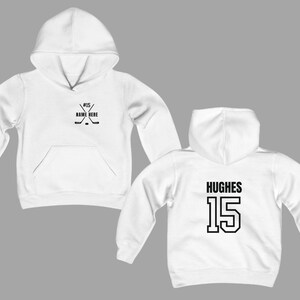 May include: White hoodie with a front pocket and hood. The front features a hockey stick graphic with the text "#15 NAME HERE". The back displays the name "HUGHES" and the number "15" in black block letters. A casual sports-themed apparel item.