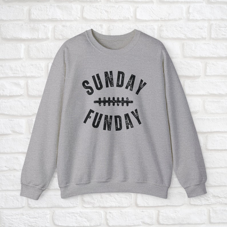 Op de afbeelding: Een grijs sweatshirt met een zwarte grafische afbeelding waarop "SUNDAY FUNDAY" staat met een voetbalnaadontwerp.