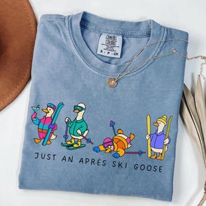 May include: A blue Comfort Colors t-shirt with a graphic of cartoon geese in ski attire and drinking cocktails. The text "JUST AN APRÈS SKI GOOSE" is printed below the geese. A gold necklace is draped across the shirt.