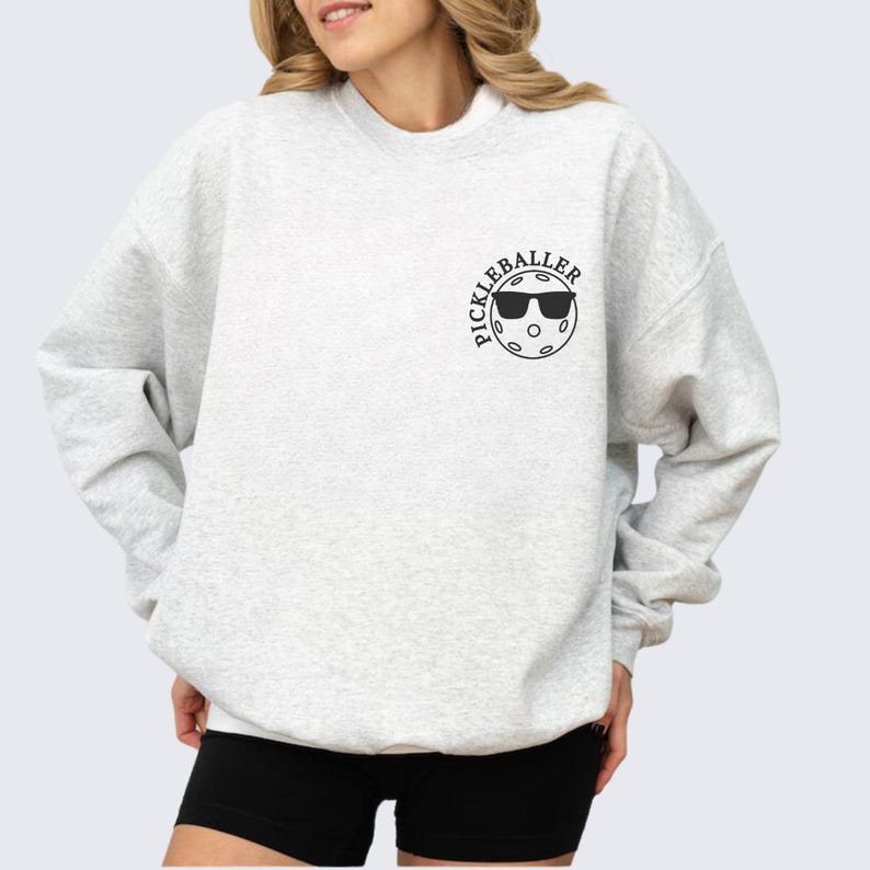 May include: A light gray sweatshirt with a black graphic of a pickleball with sunglasses and the text "Pickleballer".