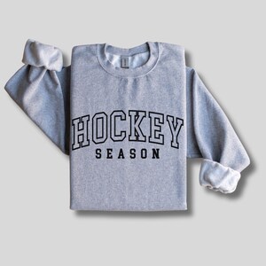 Pu&ograve; includere: Una felpa grigia con il testo "HOCKEY SEASON" in lettere nere con contorno.