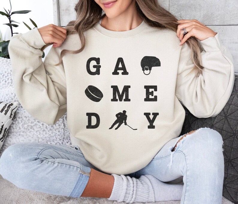 K&ouml;nnte beinhalten: Ein cremefarbener Pullover mit dem Text "GAME DAY" in schwarzen Buchstaben. Die Buchstaben sind in einem Raster angeordnet, wobei sich ein Hockeypuck, ein Hockeyspieler und ein Hockeyhelm in den Zwischenr&auml;umen befinden.