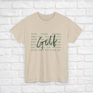 K&ouml;nnte beinhalten: Ein beiges T-Shirt mit dem Wort "Golf" in gro&szlig;en, gr&uuml;nen Buchstaben. Das Wort "Golf" ist umgeben von einer Liste von Golfbegriffen, darunter "Birdie", "Bunker", "Mulligan", "Bogey", "Chip", "Fore", "Three Putt", "Rough", "Slice", "Green", "Chunk", "Hook", "Drive", "And", "Wedge", "Pitch", "Draw", "Wing", "Shank", "Spin", "Fairway", "Iron", "Tee Time", "Putter", "Eagle", "Hole in One" und "Par".