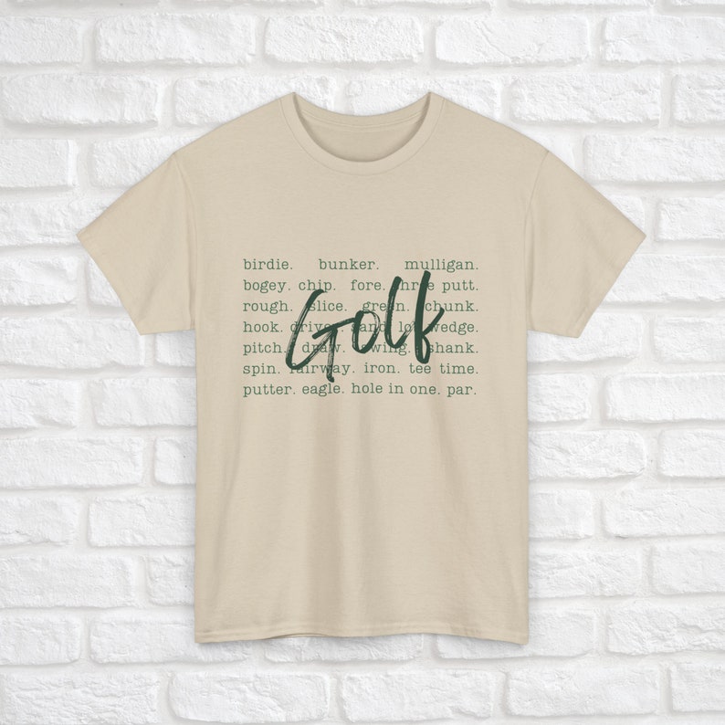 K&ouml;nnte beinhalten: Ein beiges T-Shirt mit dem Wort "Golf" in gro&szlig;en, gr&uuml;nen Buchstaben. Das Wort "Golf" ist umgeben von einer Liste von Golfbegriffen, darunter "Birdie", "Bunker", "Mulligan", "Bogey", "Chip", "Fore", "Three Putt", "Rough", "Slice", "Green", "Chunk", "Hook", "Drive", "And", "Wedge", "Pitch", "Draw", "Wing", "Shank", "Spin", "Fairway", "Iron", "Tee Time", "Putter", "Eagle", "Hole in One" und "Par".