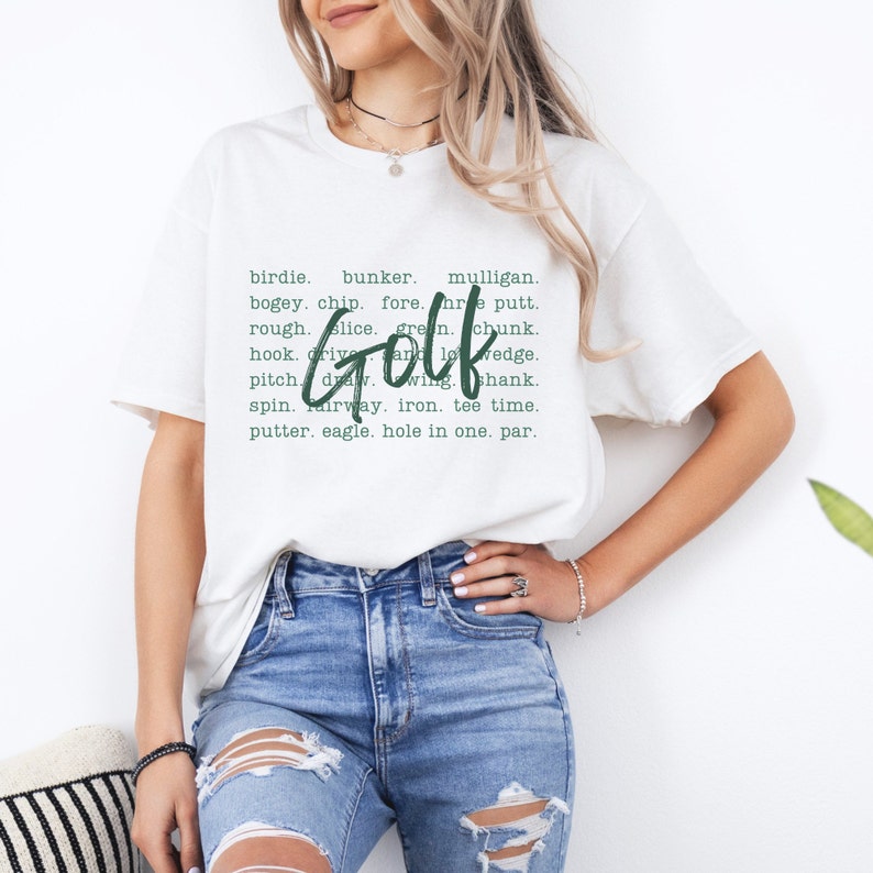K&ouml;nnte beinhalten: Wei&szlig;es T-Shirt mit einer gr&uuml;nen Grafik, auf der "Golf" steht und verschiedene Golfbegriffe aufgelistet sind, darunter Birdie, Bunker, Mulligan, Bogey, Chip, Fore, Putt, Rough, Slice, Green, Chunk, Hook, Drive, Wedge, Pitch, Draw, Wing, Shank, Spin, Fairway, Eisen, Abschlagzeit, Putter, Eagle, Hole-in-One und Par.