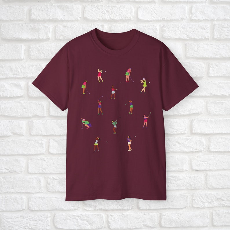 Może przedstawiać: Bordowy t-shirt z wzorem kobiet grających w golfa w r&oacute;żnych pozach. Kobiety są ubrane w r&oacute;żne kolory i trzymają kije golfowe.