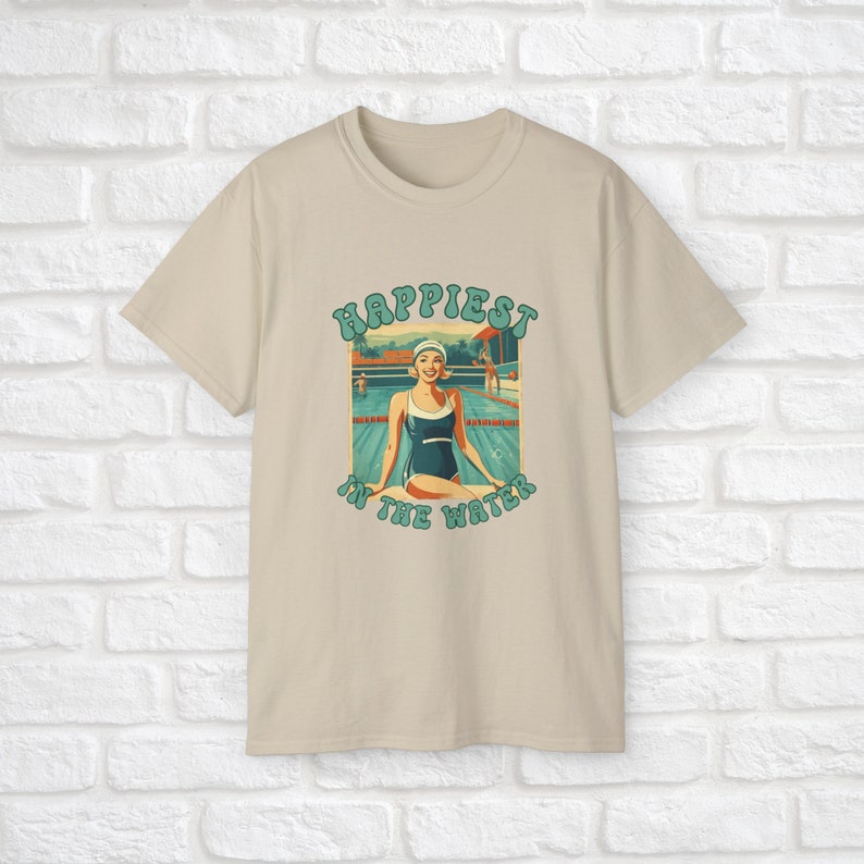 May include: A beige t-shirt with a retro-style graphic of a woman in a swimming pool. The graphic says "Happiest in the Water".