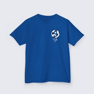 May include: Royal blue t-shirt with a white and black soccer ball graphic featuring the number 9. The shirt has a white signature that says "Taylor" underneath the ball.