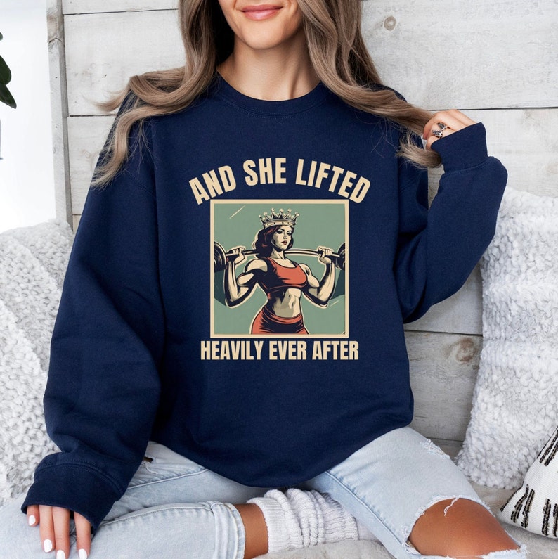 May include: Navy blue sweatshirt with a vintage-style graphic of a woman wearing a crown and lifting weights. The text reads "And She Lifted Heavily Ever After."