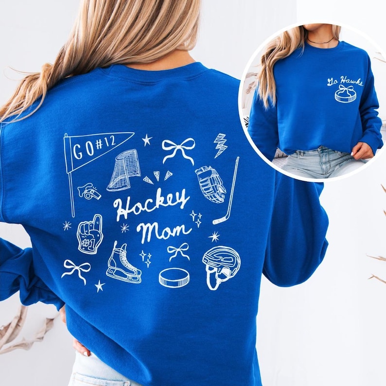 Woman wearing a crewneck sweatshirt with “Hockey Mom” doodle graphic on the back and personalized team name "Go Hawks" and player number "12" on the front; casual hockey game day outfit for moms.
