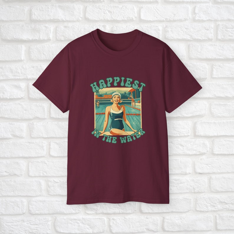 May include: A maroon t-shirt with a retro-style graphic of a woman in a swimsuit sitting by a pool. The graphic says "Happiest in the water".
