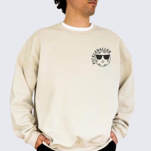 May include: A beige sweatshirt with a black graphic of a pickleball with sunglasses and the text "Pickleballer" on the front.