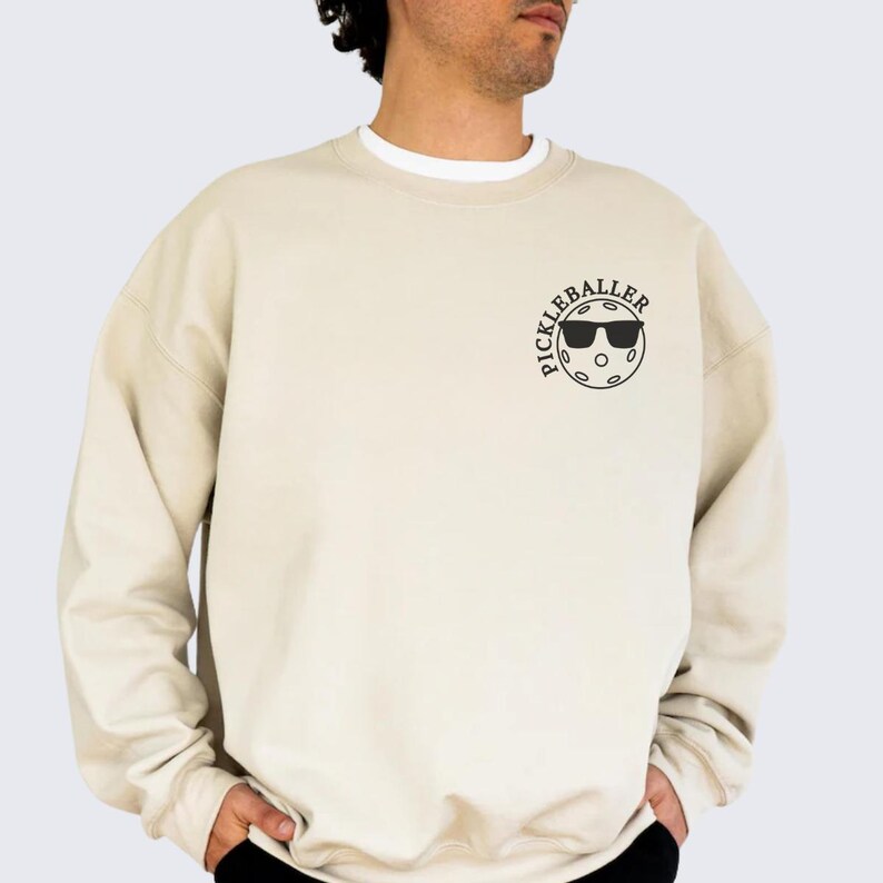 May include: A beige sweatshirt with a black graphic of a pickleball with sunglasses and the text "Pickleballer" on the front.