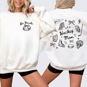 Woman wearing a crewneck sweatshirt with “Hockey Mom” doodle graphic on the back and personalized team name "Go Hawks" and player number "12" on the front; casual hockey game day outfit for moms.
