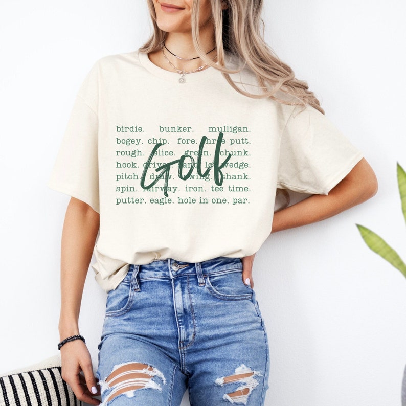 K&ouml;nnte beinhalten: Ein wei&szlig;es T-Shirt mit einer gr&uuml;nen Grafik, auf der "Golf" steht, und einer Liste von Golfbegriffen. Die Begriffe umfassen Birdie, Bunker, Mulligan, Bogey, Chip, Fore, Green, Putt, Rough, Slice, Chunk, Hook, Drive, Wedge, Pitch, Draw, Wing, Shank, Spin, Fairway, Eisen, Abschlagzeit, Putter, Eagle, Hole-in-One und Par.