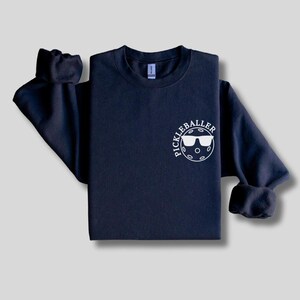 May include: A navy blue sweatshirt with a white graphic of a pickleball with sunglasses and the text "Pickleballer" on the front.