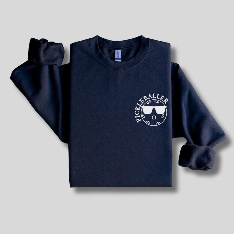 May include: A navy blue sweatshirt with a white graphic of a pickleball with sunglasses and the text "Pickleballer" on the front.