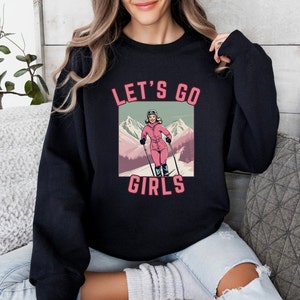 May include: Black sweatshirt featuring a graphic of a woman skiing down a mountain. The text "Let's Go Girls" is printed on the sweatshirt.