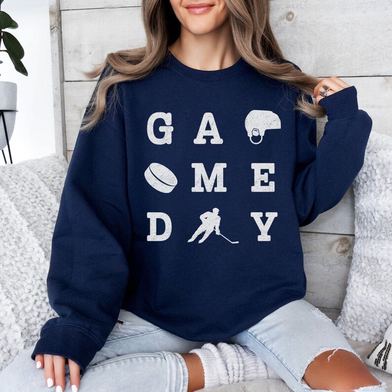K&ouml;nnte beinhalten: Ein marineblauer Pullover mit den Worten "GAME DAY" in Wei&szlig;, wobei einige Buchstaben durch Hockey-Symbole ersetzt wurden.