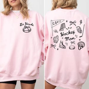 Woman wearing a crewneck sweatshirt with “Hockey Mom” doodle graphic on the back and personalized team name "Go Hawks" and player number "12" on the front; casual hockey game day outfit for moms.