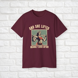 Może przedstawiać: Bordowy t-shirt z grafiką przedstawiającą kobietę w koronie podnoszącą ciężary. Na koszulce nadrukowany jest tekst "And She Lifted Heavily Ever After".