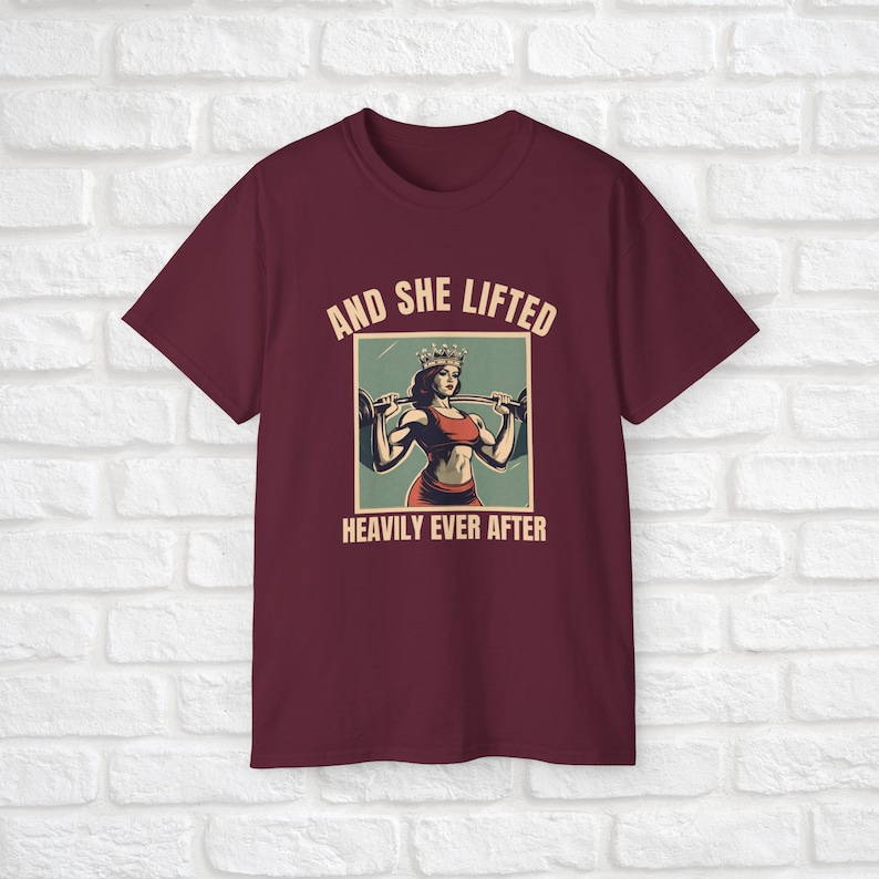 Może przedstawiać: Bordowy t-shirt z grafiką przedstawiającą kobietę w koronie podnoszącą ciężary. Na koszulce nadrukowany jest tekst "And She Lifted Heavily Ever After".