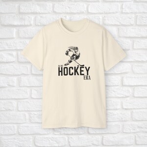 Puede incluir: Una camiseta de color crema con una ilustraci&oacute;n en blanco y negro de un jugador de hockey y el texto "In my hockey era".