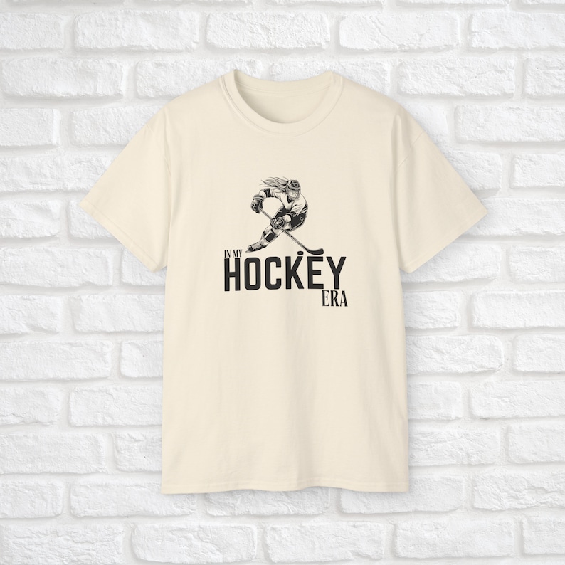 Puede incluir: Una camiseta de color crema con una ilustraci&oacute;n en blanco y negro de un jugador de hockey y el texto "In my hockey era".