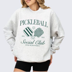 Puede incluir: Una sudadera gris con un dise&ntilde;o gr&aacute;fico verde que dice "Pickleball Social Club Est. 2025 Dink Responsibly". El gr&aacute;fico presenta dos palas de pickleball y una pelota.