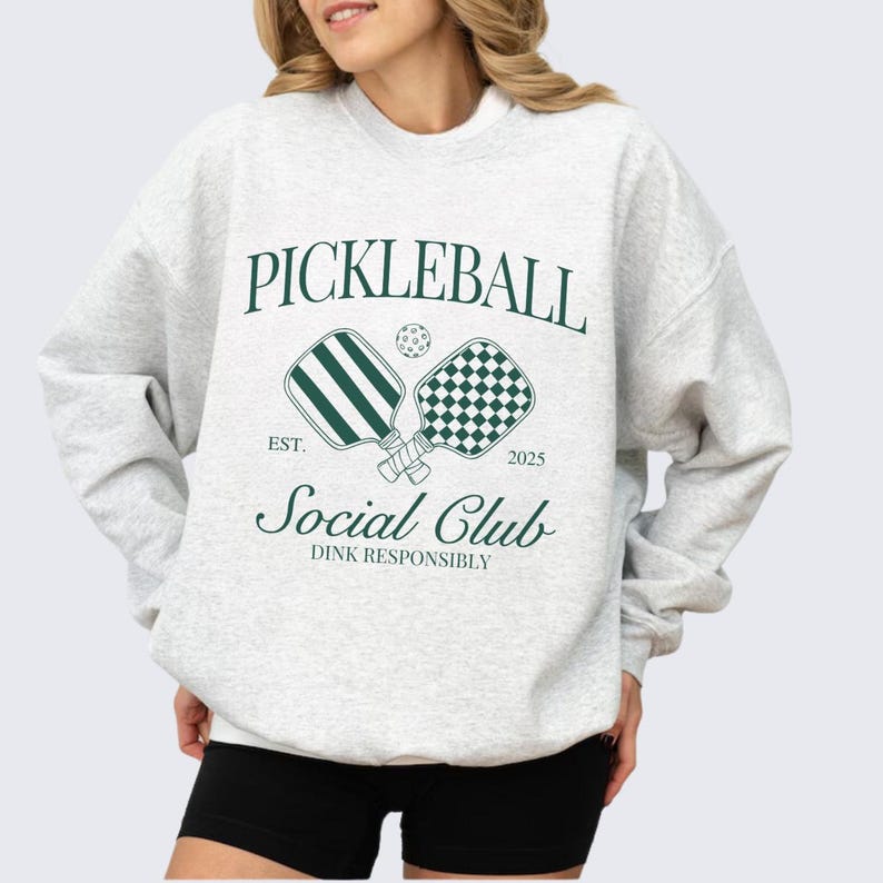 Puede incluir: Una sudadera gris con un dise&ntilde;o gr&aacute;fico verde que dice "Pickleball Social Club Est. 2025 Dink Responsibly". El gr&aacute;fico presenta dos palas de pickleball y una pelota.