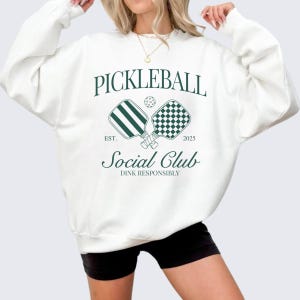 Puede incluir: Sudadera blanca con un dise&ntilde;o gr&aacute;fico verde que dice "Pickleball Social Club Est. 2025 Dink Responsibly". El gr&aacute;fico presenta dos palas de pickleball cruzadas y una pelota.