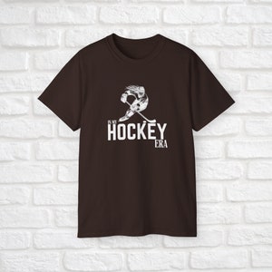 Puede incluir: Una camiseta marr&oacute;n con un gr&aacute;fico blanco de un jugador de hockey patinando y sosteniendo un palo. El texto "In My Hockey Era" est&aacute; impreso debajo del gr&aacute;fico.