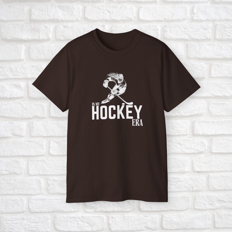 Puede incluir: Una camiseta marr&oacute;n con un gr&aacute;fico blanco de un jugador de hockey patinando y sosteniendo un palo. El texto "In My Hockey Era" est&aacute; impreso debajo del gr&aacute;fico.