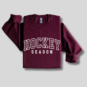 Pu&ograve; includere: Una felpa color bordeaux con testo bianco che recita "HOCKEY SEASON".