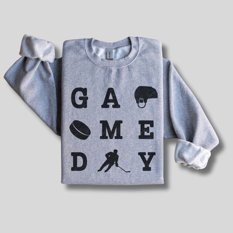 K&ouml;nnte beinhalten: Ein grauer Pullover mit dem Aufdruck "GAME DAY" in Schwarz. Die Buchstaben sind von Hockey-Motiven umgeben, darunter ein Puck, ein Schl&auml;ger und ein Helm.