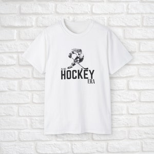 Puede incluir: Camiseta blanca con una ilustraci&oacute;n en blanco y negro de un jugador de hockey y el texto "In My Hockey Era".