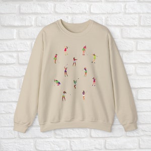 May include: A beige sweatshirt featuring a pattern of women playing golf. The women are wearing different coloured clothing and are in various poses.