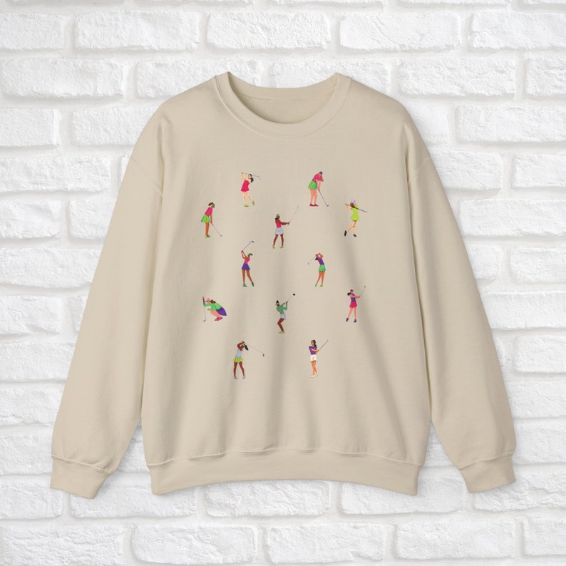 May include: A beige sweatshirt featuring a pattern of women playing golf. The women are wearing different coloured clothing and are in various poses.