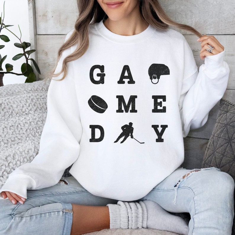 K&ouml;nnte beinhalten: Wei&szlig;er Pullover mit den Worten "GAME DAY" in schwarzen Buchstaben. Die Buchstaben sind in einem Raster angeordnet, mit einem Hockeypuck, einem Hockeyspieler und einem Hockeyhelm in den Zwischenr&auml;umen zwischen den Buchstaben.