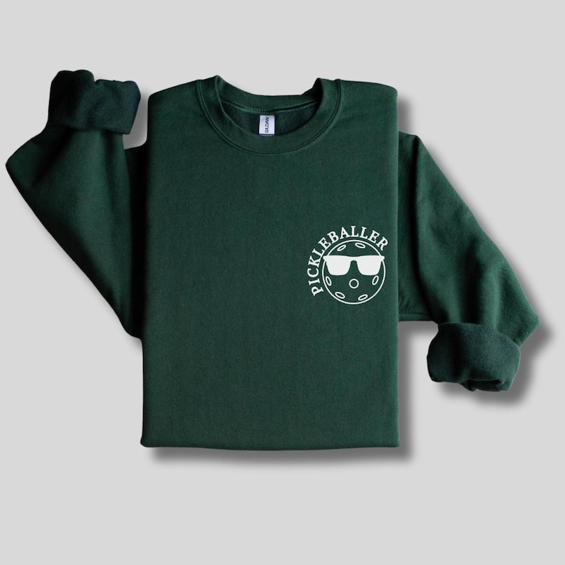 May include: A dark green sweatshirt with a white circle graphic that says "Pickleballer" and features a pickleball with sunglasses.