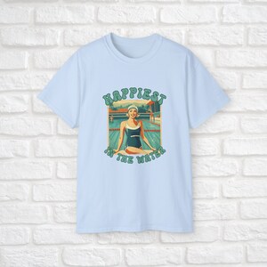 May include: Light blue t-shirt with a retro-style graphic of a woman in a swimsuit sitting by a pool. The graphic says "Happiest in the water".
