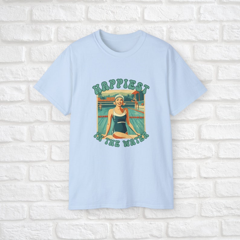 May include: Light blue t-shirt with a retro-style graphic of a woman in a swimsuit sitting by a pool. The graphic says "Happiest in the water".