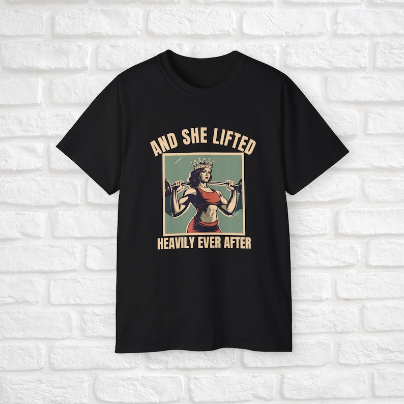 Może przedstawiać: Czarny T-shirt z grafiką w stylu vintage przedstawiającą kobietę w koronie podnoszącą ciężary. Tekst brzmi "And she lifted heavily ever after."
