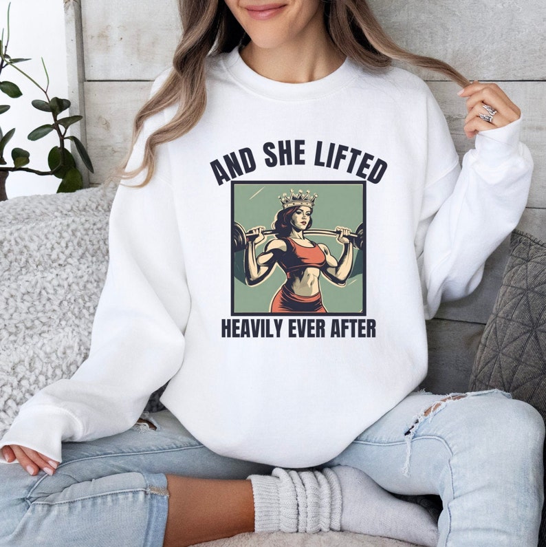 May include: White sweatshirt with a graphic of a woman wearing a crown and lifting weights. The text reads "And she lifted heavily ever after."