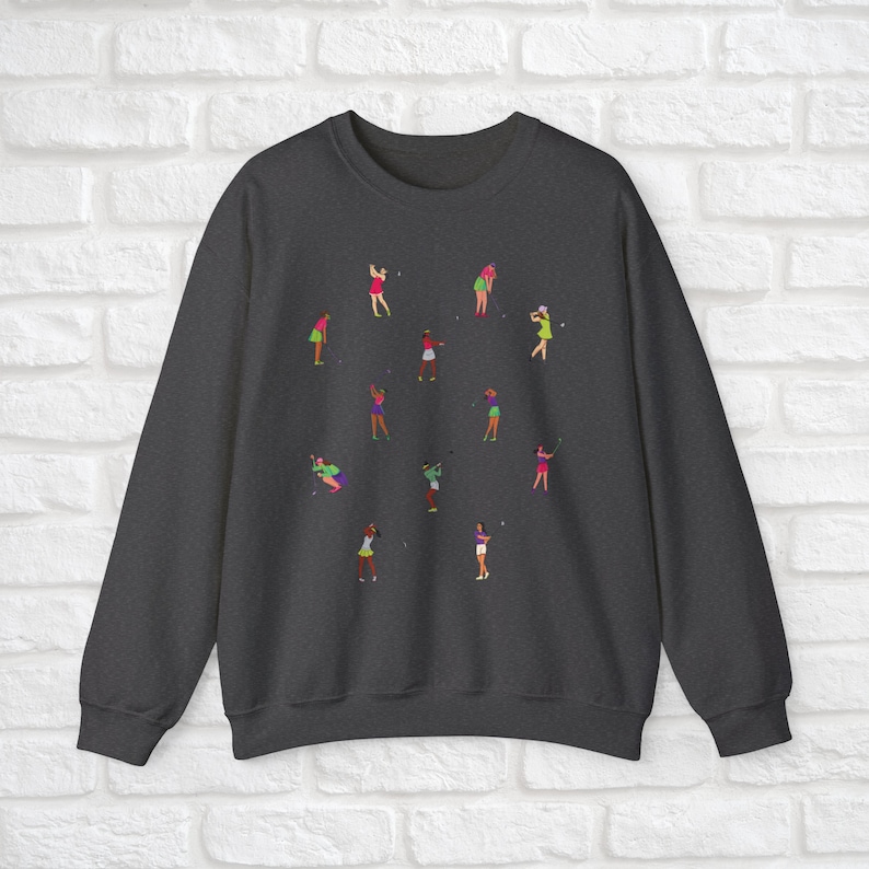 May include: Dark grey sweatshirt featuring a design of women playing golf in different poses. The women are wearing pink, green and white clothing.